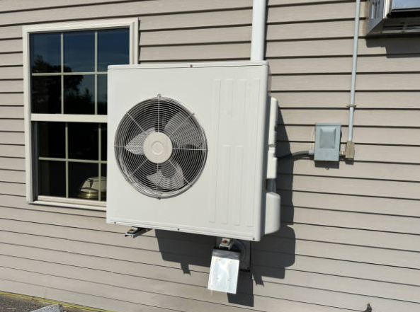 An outdoor HVAC condenser unit is mounted on the exterior wall of a home next to a window.