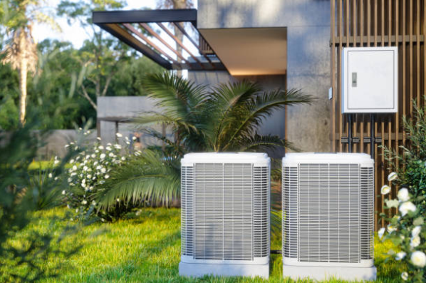 Two outdoor HVAC condenser units are installed beside a modern home surrounded by landscaping and greenery.