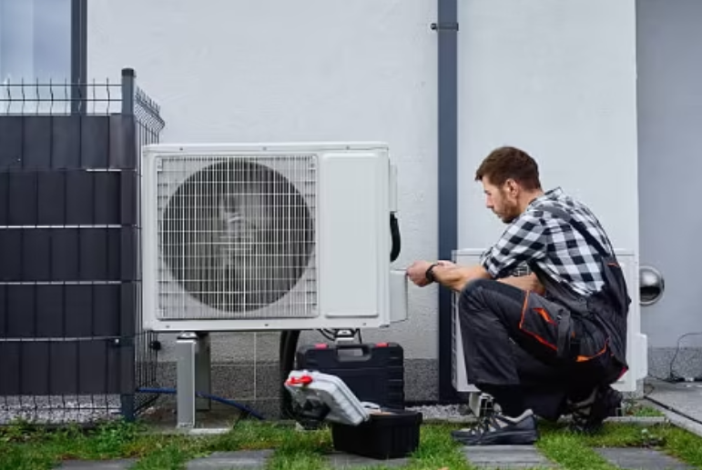 A technician crouches outdoors while repairing an air conditioning unit with tools nearby.