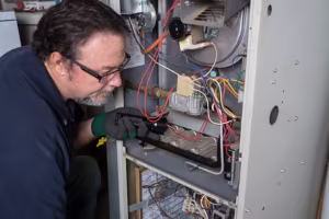 Technician using a flashlight to closely inspect the wiring and burner assembly inside an open furnace unit.