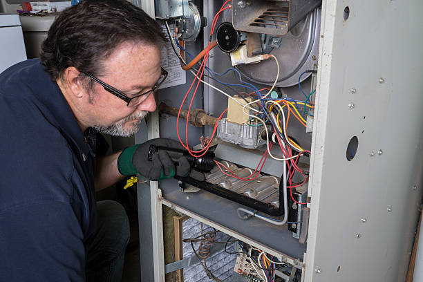 Technician using a flashlight to closely inspect the wiring and burner assembly inside an open furnace unit.