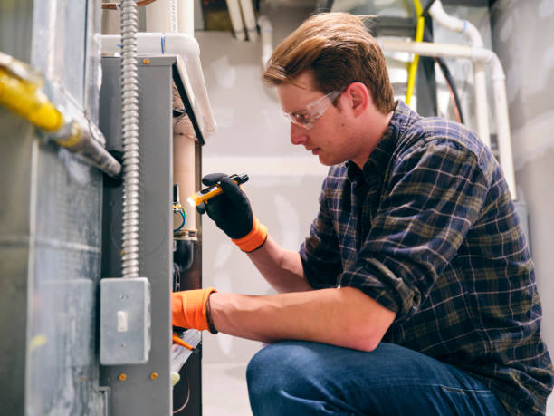 Technician wearing safety glasses and gloves crouching to inspect a furnace unit with a flashlight in a utility room.