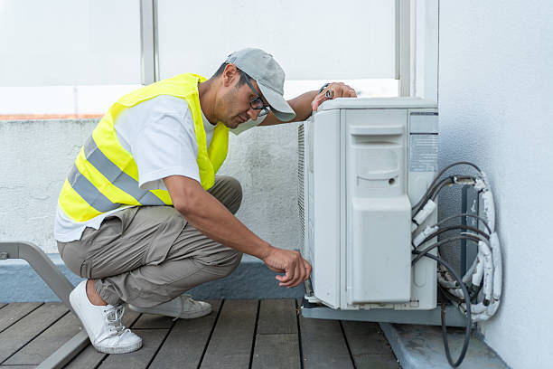Technician wearing a yellow safety vest crouching to inspect an outdoor air conditioning unit on a rooftop deck.