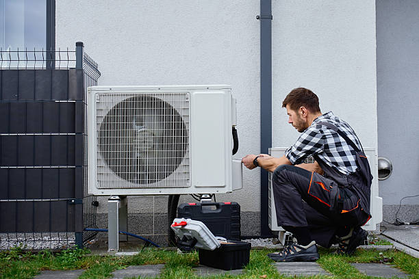 Technician crouching beside an open toolbox while servicing an outdoor air conditioning condenser unit next to a house.