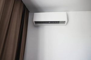 White wall-mounted indoor air conditioning unit installed in the corner of a room near a curtained window.